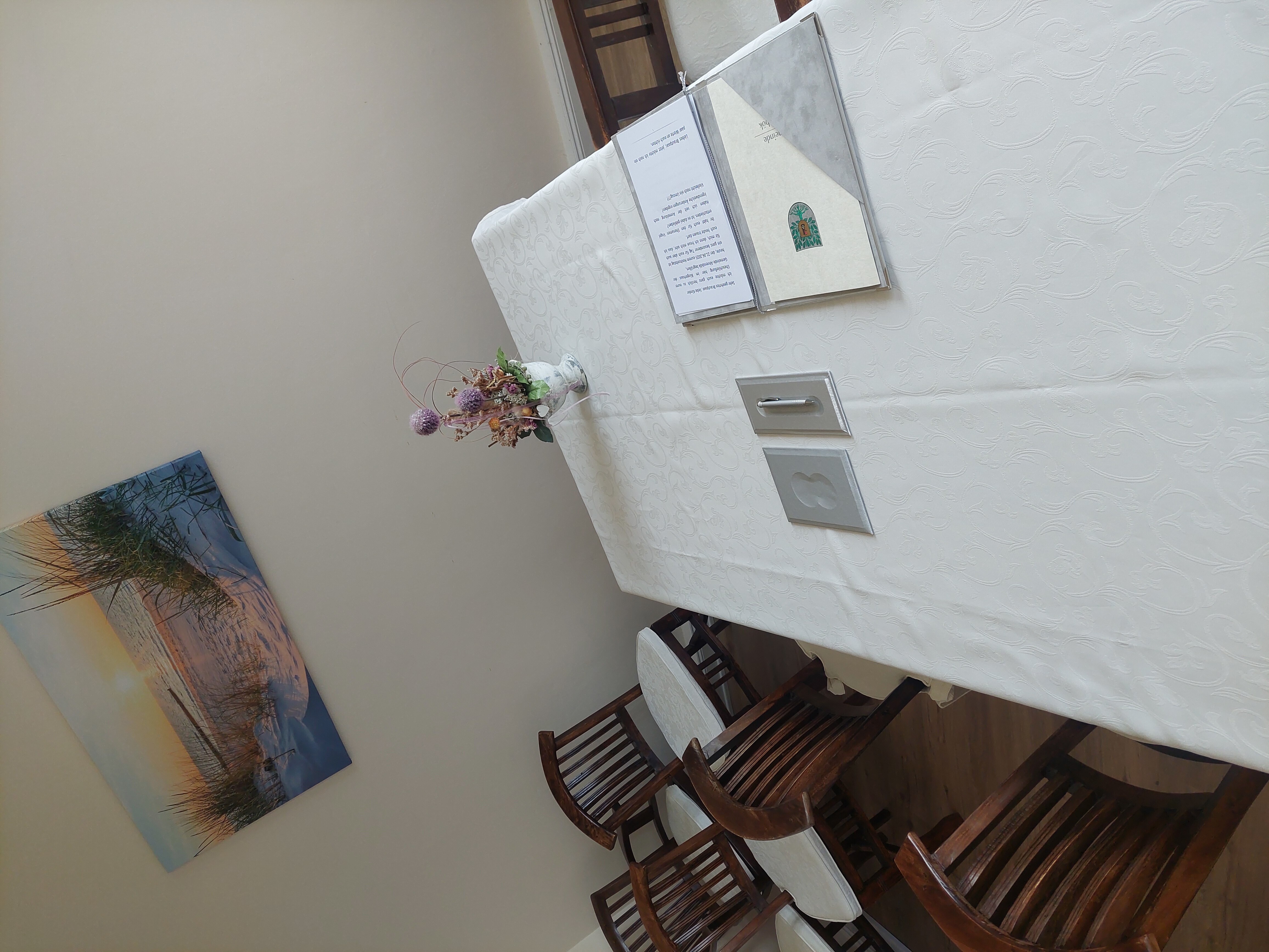 Bild vom Trauzimmer im Bürgerhaus mit braunen Stühlen und einem Tisch mit heller Tischdecke auf dem ein Ordner liegt und eine Blume steht. An der Wand hängt ein Strandbild vom Sonnenuntergang. Bild vom Trauzimmer im Bürgerhaus mit braunen Stühlen und einem Tisch mit heller Tischdecke auf dem ein Ordner liegt und eine Blume steht. An der Wand hängt ein Strandbild vom Sonnenuntergang.